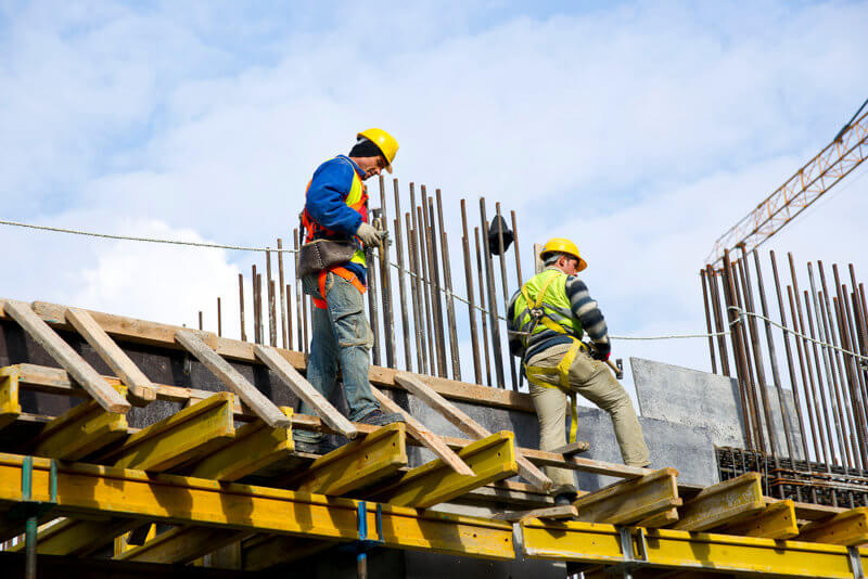 Two construction workers wearing hard hats and safety vests stand on wooden scaffolding beside exposed vertical rebar at a building site under a blue sky, formwork in progress.