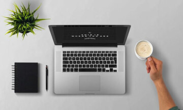 Overhead view of a laptop centered on a gray desk with a notebook and pen to the left, a plant in the top-left, and a hand reaching for a cup of coffee on the right.