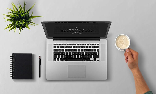 Smart_working - S.T.A. Barbotta Overhead view of a laptop centered on a gray desk with a notebook and pen to the left, a plant in the top-left, and a hand reaching for a cup of coffee on the right.